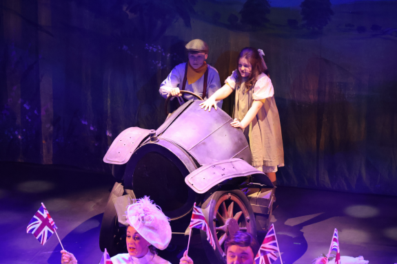 A man and a girl interact with a vintage car on a theatrical stage with scenic backdrop.