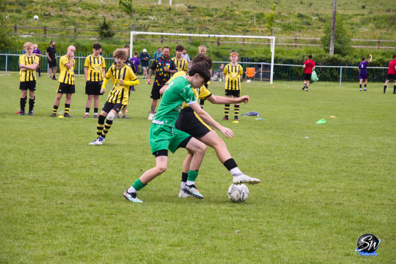Two children playing football, one in green tackling the other in black, with teams in the background.