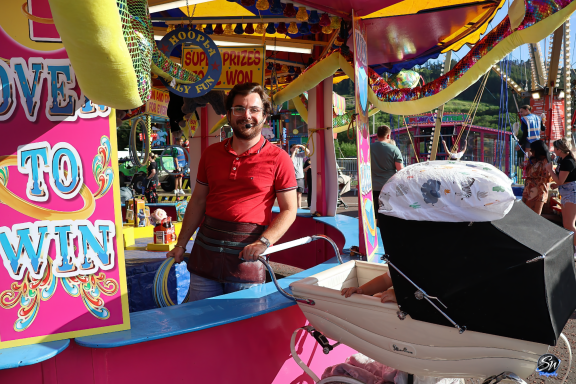 A man in a red shirt stands by a colourful carnival game booth with a baby stroller nearby.