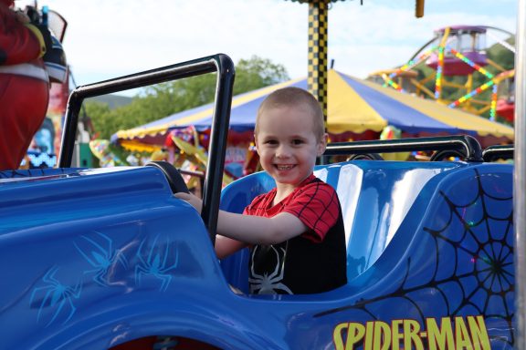 A young boy in a Spider-Man shirt smiles while sitting in a ride at a fairground.