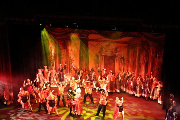 A vibrant stage performance with dancers and performers in costume, illuminated by colorful lights.