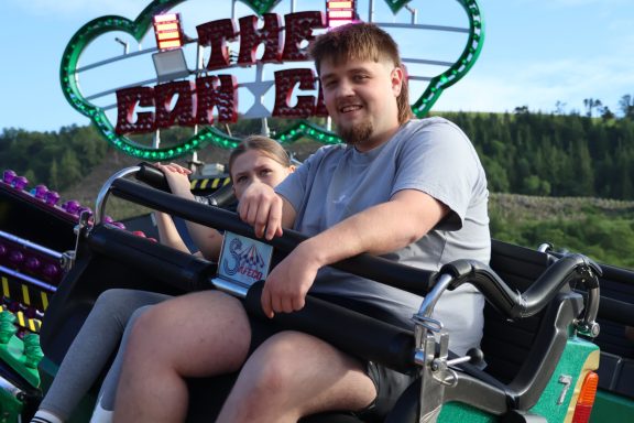 Two people sit in a ride at an amusement park, smiling in front of vibrant signs and greenery.
