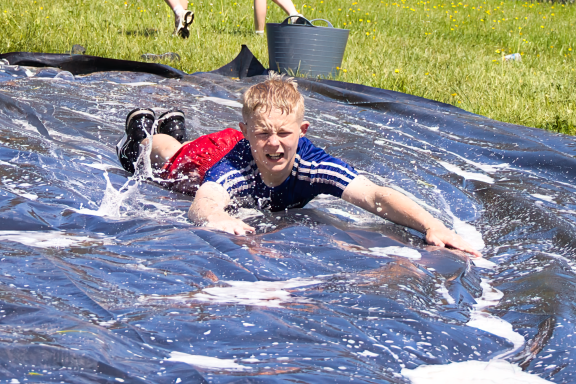 A boy in a blue shirt slides on a slip-n-slide, water splashing around him.