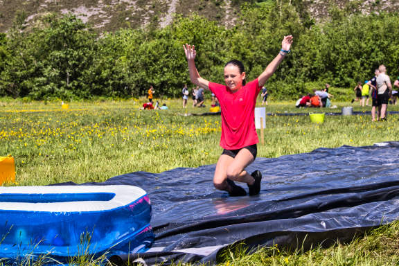 A child joyfully slides on a water slide in a grassy field with people in the background.