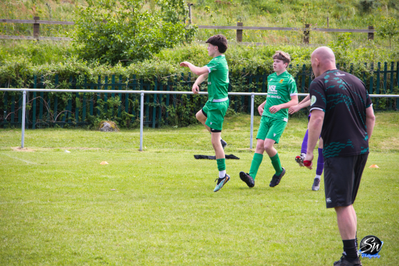 Two players in green football kits practice on a grassy field, while a coach observes.