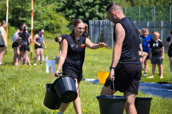 Two participants carrying buckets during an outdoor activity in a grassy field.