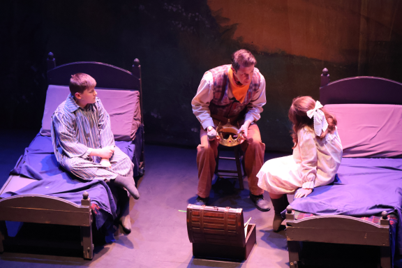 Two beds with actors sitting and one standing between them, in a theatrical setting.