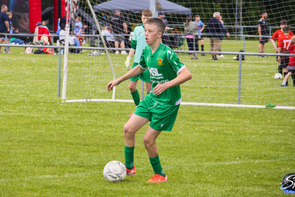 A young male footballer in a green kit prepares to kick a ball on a grassy field.