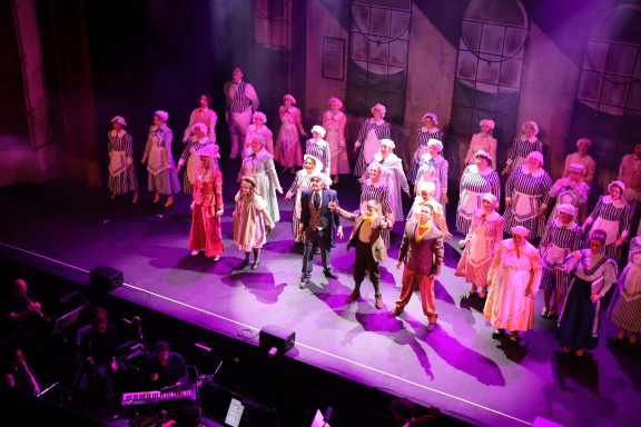 A large musical theatre cast performing on stage with vibrant costumes and dramatic lighting.