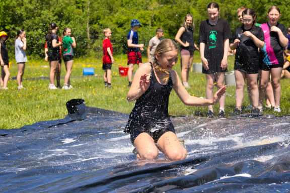 A girl slides on a water-slicked surface while others watch and enjoy on a sunny day.