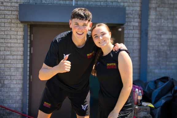 Two smiling young people pose for a photo, one giving a thumbs up, both in sports attire.