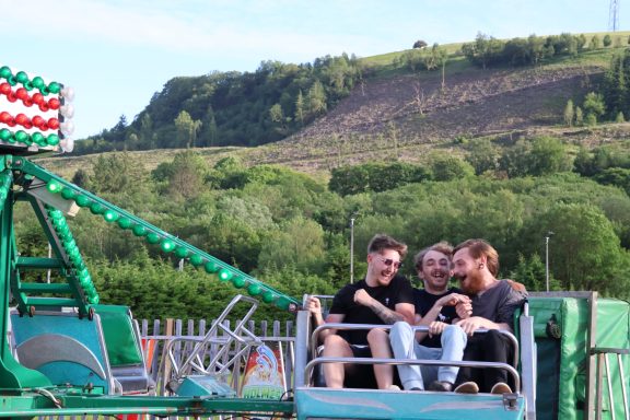 Three young men laughing together on a fairground ride, with green hills in the background.