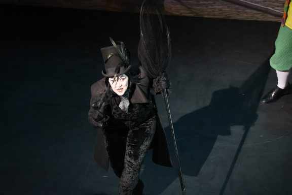 A performer in a dark costume and top hat, holding a staff, looks up against a dim backdrop.