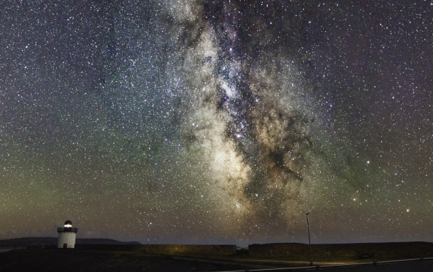 A panoramic view of the Milky Way with a lighthouse in the foreground.