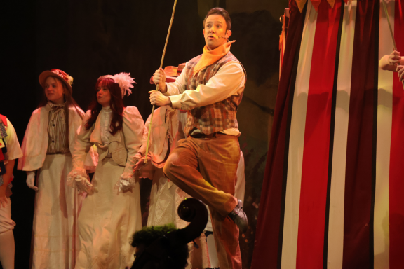 A performer in costume swings from a rope while others dressed in period attire watch.