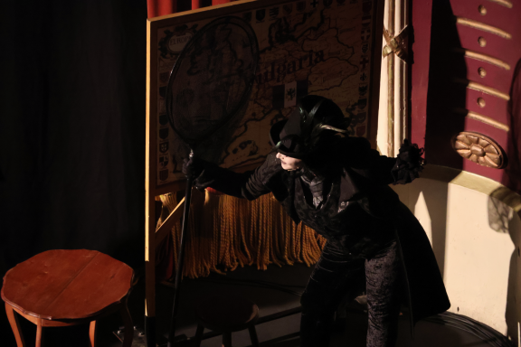 A performer in a dark costume mimics an animal on stage, shadowed by a backdrop.