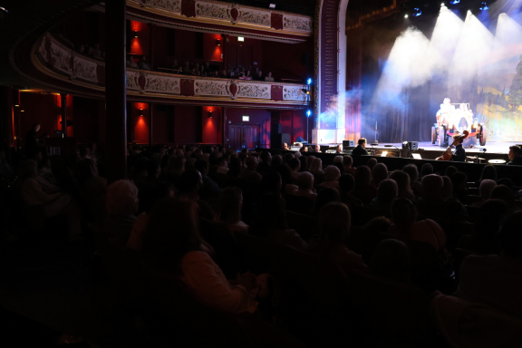 Audience in a theatre enjoying a performance with stage lights illuminating the scene.