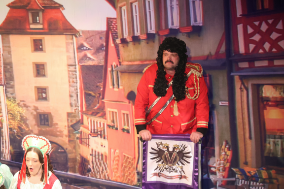 Man in a red coat with black fur trim stands, holding a banner, in a festive setting.