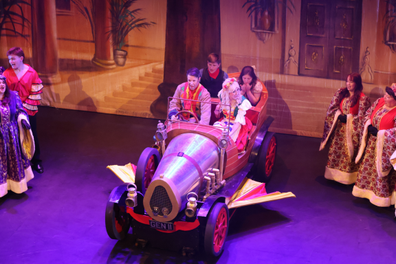 A vintage car on stage with performers in costumes celebrating a theatrical scene.