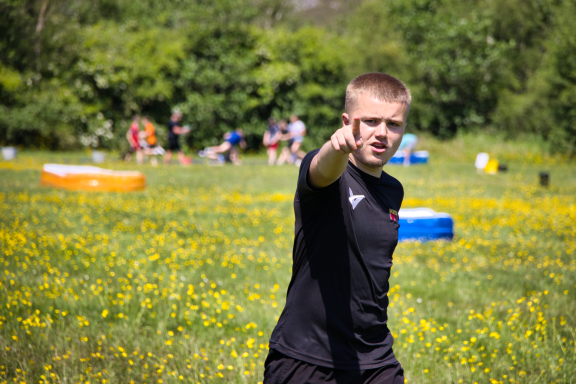 A young man in a black shirt points while standing in a sunny field with people in the background.