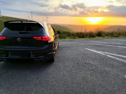 Black car parked on a road with a sunset in the background over green hills.