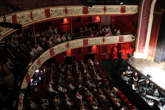 Interior of a theatre showing an audience in the balcony and a stage below.
