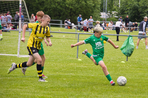 A child in green kicking a football while a player in yellow approaches.