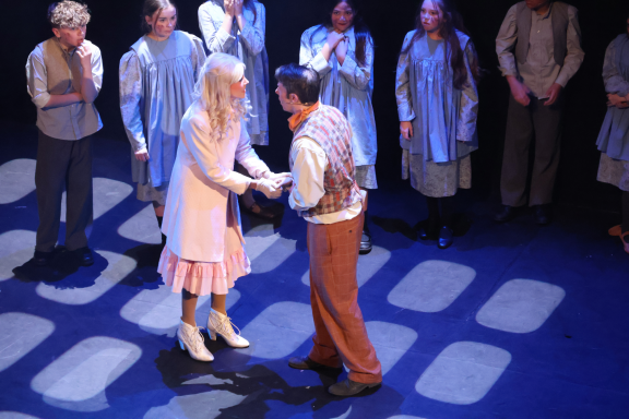 A man and woman shake hands on stage, surrounded by other performers in the background.