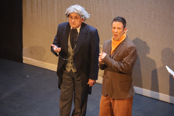 Two actors performing on stage, one in a suit and the other in a casual outfit.