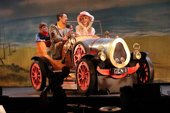 A vintage-style car with three characters, two adults and a child, dressed in period costumes.