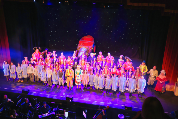 A large group of performers in costumes on stage, with colourful backgrounds and lighting.