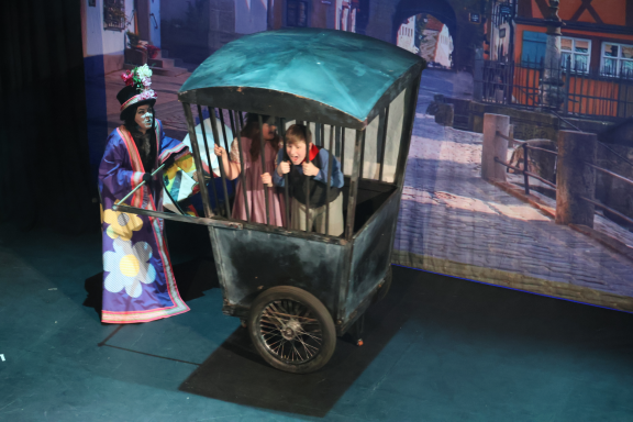 A performer in costume interacts with two people in a cage-like cart on stage.