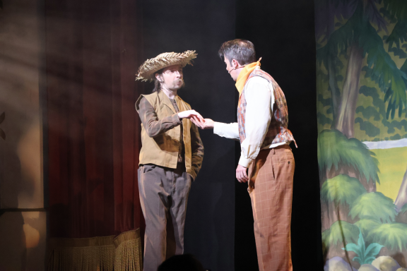 Two actors performing on stage, one wearing a cowboy hat and costume, with a scenic background.
