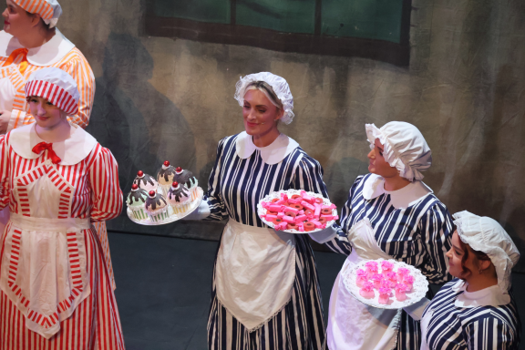 Group of women in period costumes, holding trays of desserts on a stage.