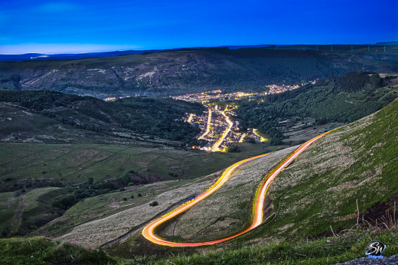 Winding road through a valley at twilight, with distant lights of a small town visible.