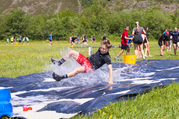 A person in black shirt and red shorts slides on a water tarp in a grassy field.