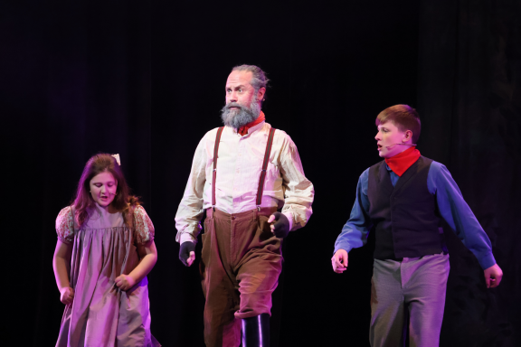 Three performers on stage: a man with a beard and two children, all in period costumes.