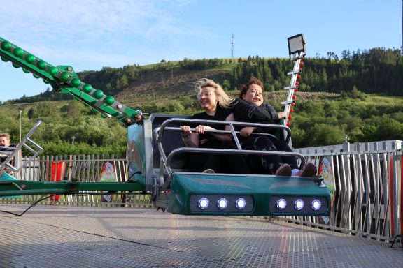 Two people enjoy a spin on a fairground ride against a green hillside backdrop.