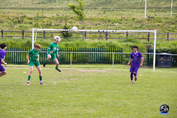 Young players in green and purple uniforms compete in a football match on a grassy field.