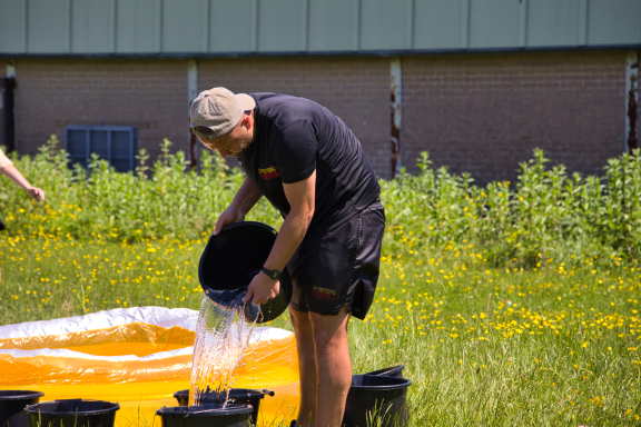 Person pouring water from a bucket onto the ground in a grassy area.