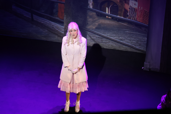 A woman in a light pink dress stands on stage, smiling under soft lighting.