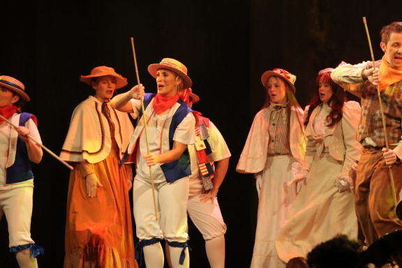 Group of performers in colourful costumes, dancing and holding props on stage.