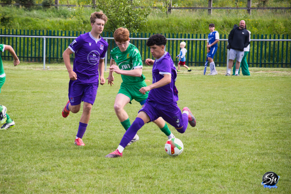 Two players in purple kits and one in green competing for a football on a grassy pitch.