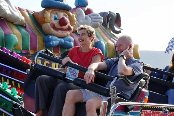 Two people smiling on a ride at a funfair, with a clown figure in the background.