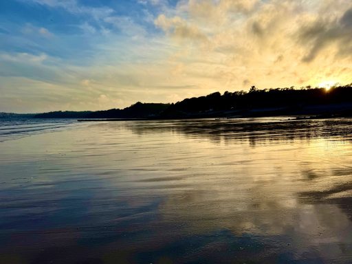 Calm beach at sunset with reflective water and silhouetted shoreline.