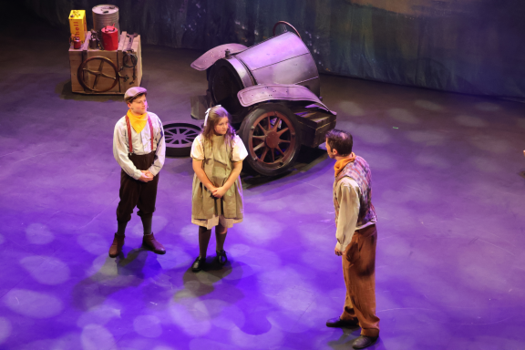 Three performers on stage with a vintage vehicle in the background and soft, coloured lights.