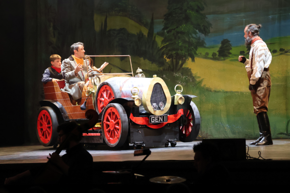 Two men in vintage clothing sit in a classic car on stage, while another man stands nearby.