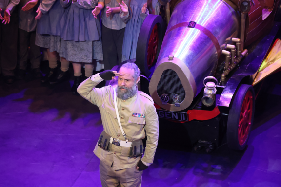 A man in a military uniform salutes in front of a whimsical cannon and a choir.