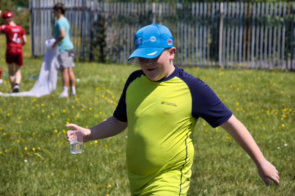 Child in a blue cap and green shirt walking on grass, holding a water bottle.