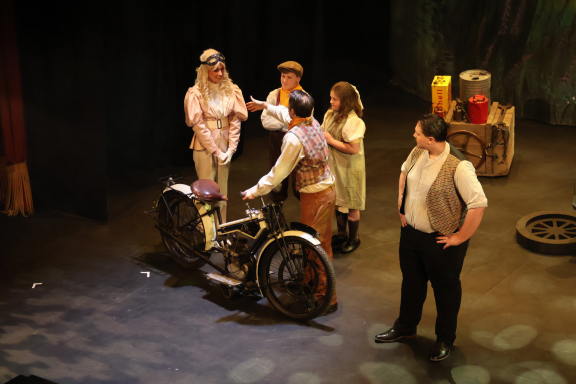 A group of five characters in period costumes interacting on stage with a bicycle.